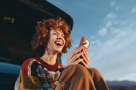 Улыбающаяся женщина с кудрявыми волосами держит смартфон для селфи рядом с припаркованной машиной на улице, жизненный портрет, яркий дневной свет, голубое небо с облаками, беззаботная свобода…
