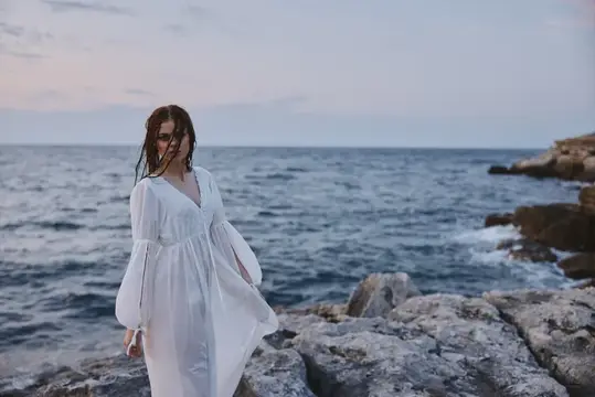 Девушка в белом платье стоит на скалах у моря