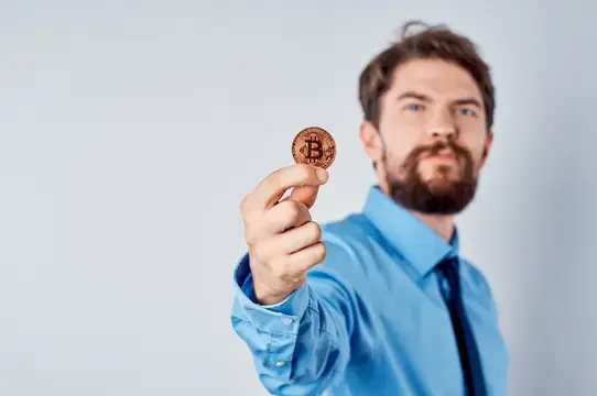 Бородатый мужчина в голубой рубашке держит биткоин на светлом фоне