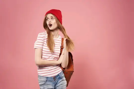 Удивлённая девушка в красной шапке с рюкзаком на розовом фоне