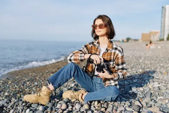Молодая женщина в клетчатой рубашке сидит с фотокамерой на галечном пляже