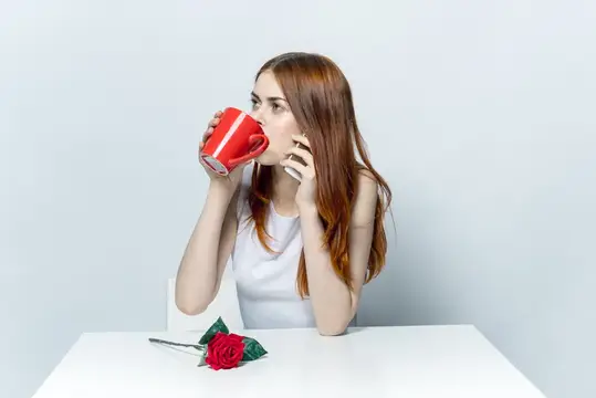 Элегантная женщина сидит за столом, пьёт из красной кружки и разговаривает по телефону