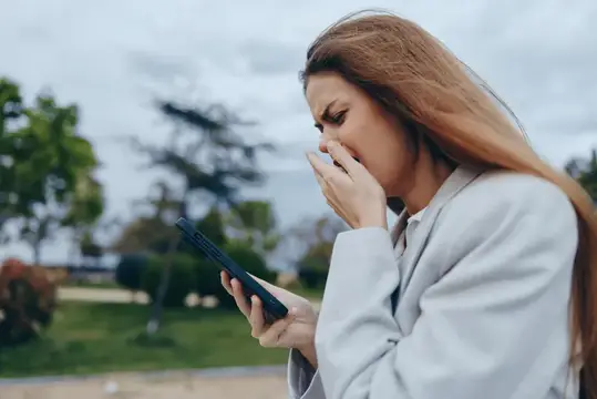 Удивлённая женщина в светлом пиджаке смотрит в телефон на улице