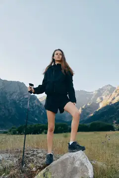 Молодая женщина с треккинговой палкой стоит на камне на фоне гор