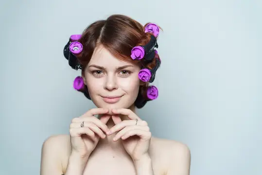 Молодая женщина с бигудями на голове улыбается на сером фоне
