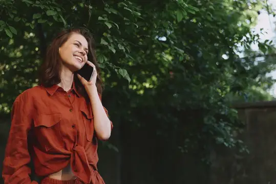 Улыбающаяся темноволосая девушка в красной рубашке говорит по телефону в парке