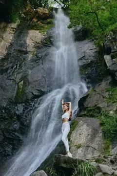 Девушка в белом спортивном костюме стоит на камнях у подножия водопада
