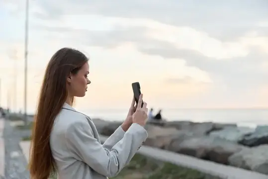 Молодая женщина в легкой куртке использует смартфон, чтобы сделать фото на открытом воздухе возле скалистого побережья во время заката, запечатлевая момент со спокойным выражением лица в мягком…