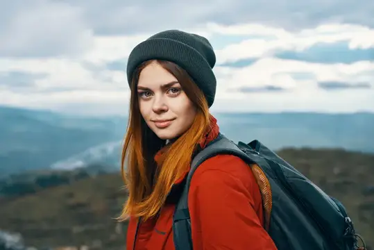 Рыжеволосая девушка в шапке с рюкзаком на фоне гор