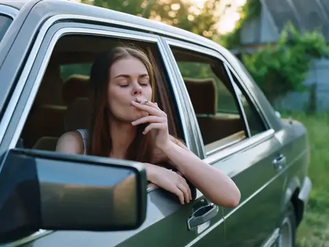 Молодая женщина с закрытыми глазами курит у окна автомобиля