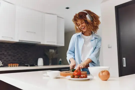 Улыбающаяся кудрявая женщина в наушниках режет овощи на светлой кухне