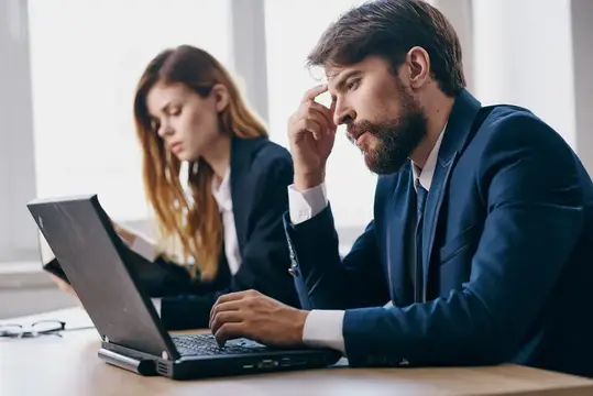 Уставший бородатый мужчина в костюме работает за ноутбуком в офисе