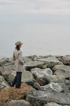 Женщина в плаще и кепке стоит на камнях у моря