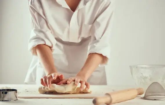 Повар в белой рубашке раскатывает тесто на кухонном столе