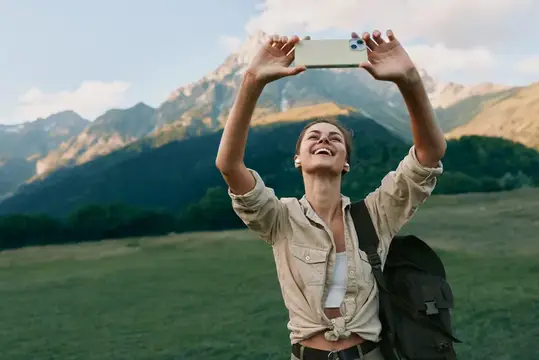 Улыбающаяся девушка с рюкзаком фотографирует горы на телефон