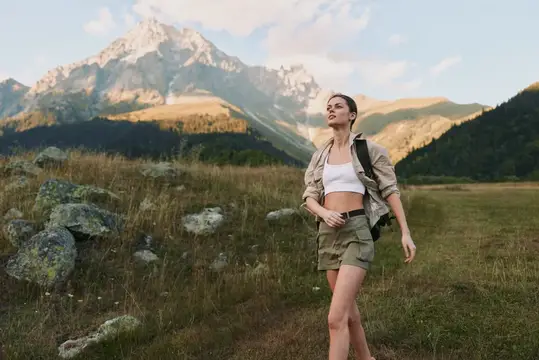 Активная женщина в походе в естественной среде, одетая в шорты и куртку, с рюкзаком, идет по горной тропе среди скалистых холмов под ясным небом