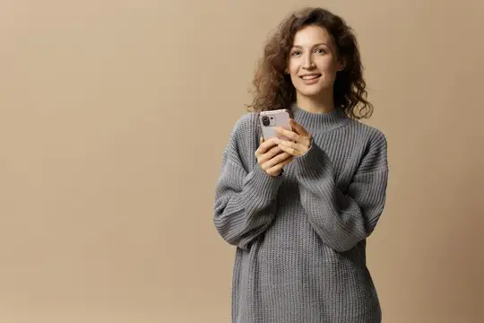 Улыбающаяся кудрявая девушка в сером свитере держит смартфон