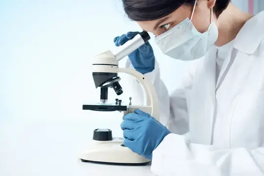 Женщина-учёный в маске и перчатках смотрит в микроскоп в лаборатории