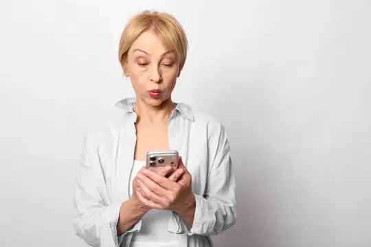 Блондинка в белой рубашке удивлённо смотрит на смартфон