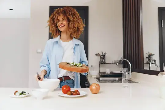 Улыбающаяся кудрявая женщина готовит салат на светлой кухне