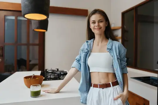 Улыбающаяся девушка в джинсовой рубашке стоит на светлой кухне