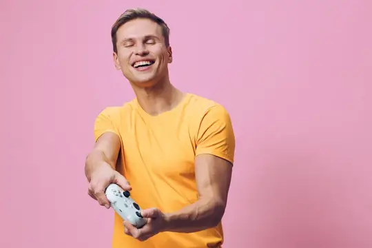 Смеющийся мужчина в желтой футболке играет в геймпад на розовом фоне