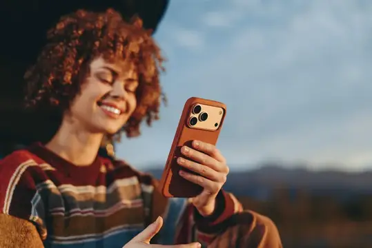 Женщина с вьющимися волосами улыбается на улице, держа смартфон для портретного селфи