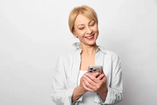 Улыбающаяся пожилая женщина в студии держит смартфон с видимым экраном, читая сообщение в чате