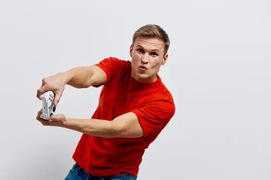 Молодой блондин в красной футболке играет в геймпад на белом фоне