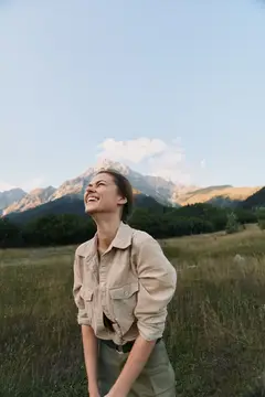 Смеющаяся девушка в бежевой куртке на фоне гор и поля