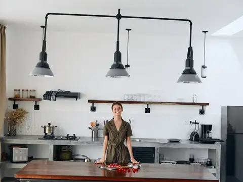 Улыбающаяся женщина стоит за деревянным столом на светлой кухне с чёрными лампами
