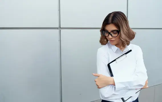 Молодая женщина в очках с планшетом на фоне серой стены