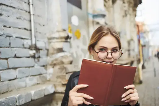 Молодая женщина в очках с красной книгой на фоне кирпичной стены