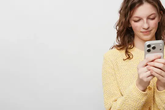 Улыбающаяся молодая женщина в желтом свитере пользуется смартфоном, стоя на белом фоне