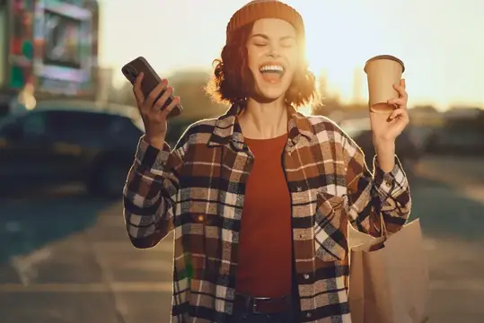 Счастливая девушка в клетчатой рубашке с телефоном и кофе на закате