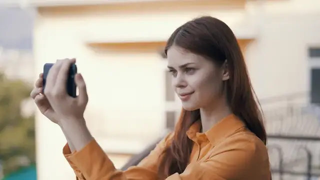 Молодая женщина в оранжевой рубашке фотографирует на смартфон на улице