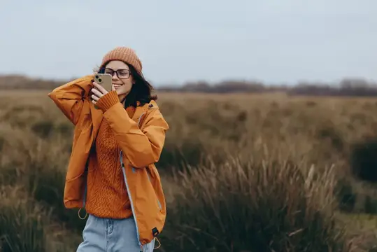 Улыбающаяся девушка в оранжевой куртке фотографирует на телефон в поле