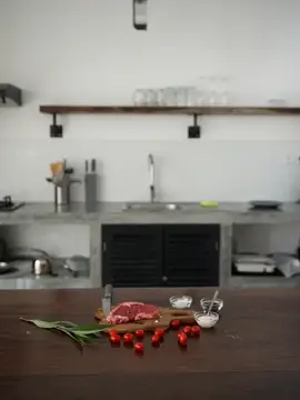 Стильная кухня, на деревянном столе лежит кусок свежего мяса со специями, помидоры и зелень, минималистичный интерьер