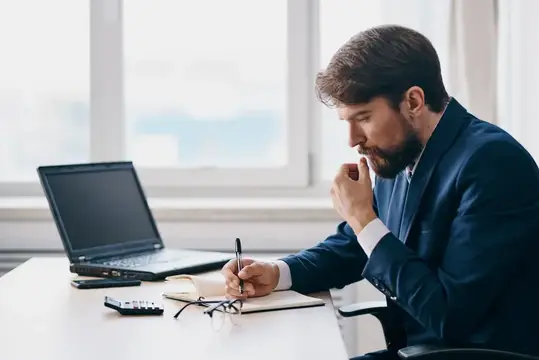 Бородатый мужчина в костюме задумчиво работает за ноутбуком в офисе