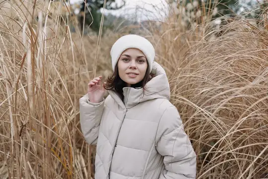 Молодая женщина в бежевом пуховике и белой шапке среди сухой травы
