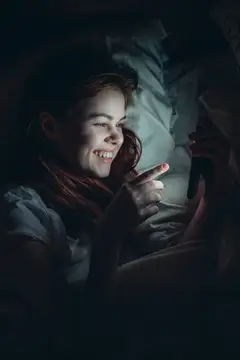 Рыжеволосая девушка улыбается, глядя на экран телефона в темноте