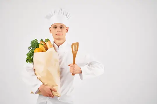 Повар в белом колпаке держит пакет с овощами и деревянную лопатку