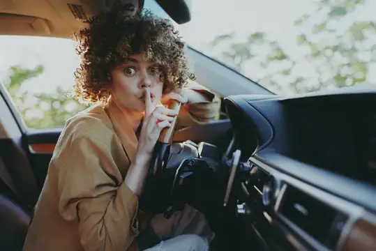 Кудрявая девушка прикладывает палец к губам в салоне автомобиля