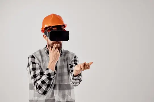 Бородатый мужчина в каске и VR-очках жестикулирует на сером фоне