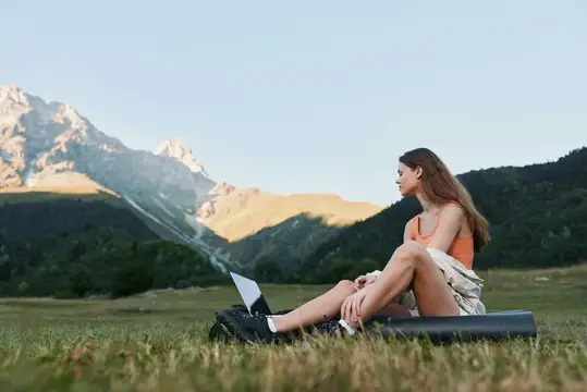 Outdoor-сцена показывает женщину, отдыхающую с ноутбуком на лугу, на фоне возвышающихся гор
