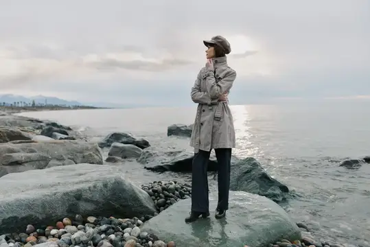 Женщина в плаще и кепке стоит на камнях у моря