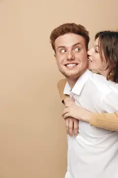 Девушка обнимает улыбающегося парня и шепчет ему на ухо на бежевом фоне