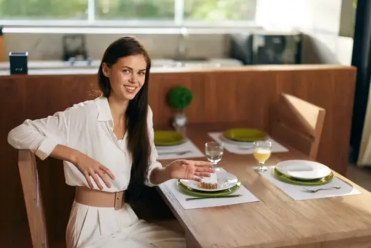 Улыбающаяся брюнетка в белом платье сидит за сервированным столом на кухне