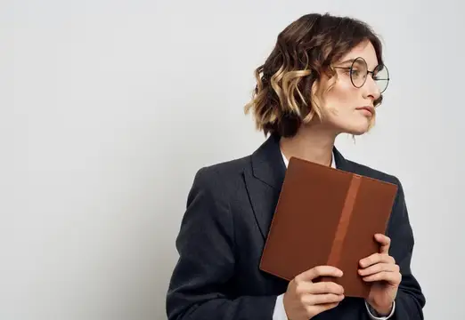 Женщина в классическом костюме с открытой книгой и очками на лице, обрезанный вид на светлом фоне
