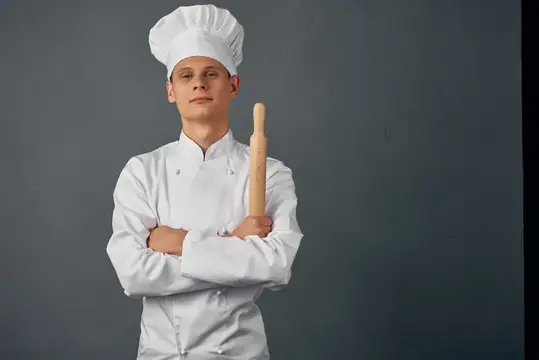 Молодой повар в белом колпаке с скалкой на сером фоне